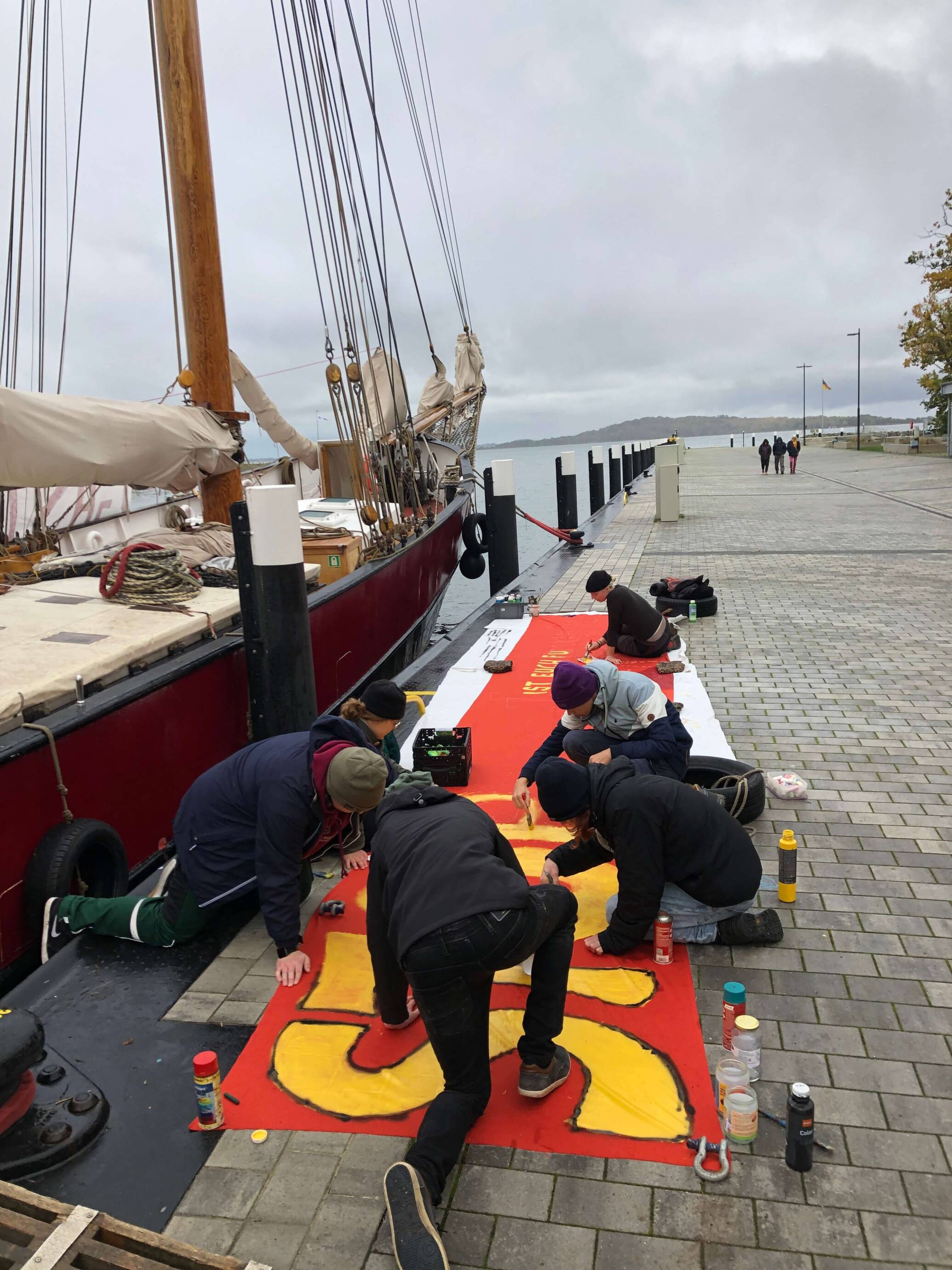 Menschen malen Banner neben Segelboot am Hafen.
